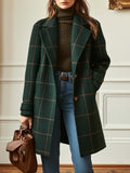Front view of a woman wearing a dark green plaid tweed mid-length coat with brown windowpane checks, wide notch lapels, and tortoiseshell front buttons, styled over an olive green turtleneck sweater, blue jeans, and holding a brown leather bag.