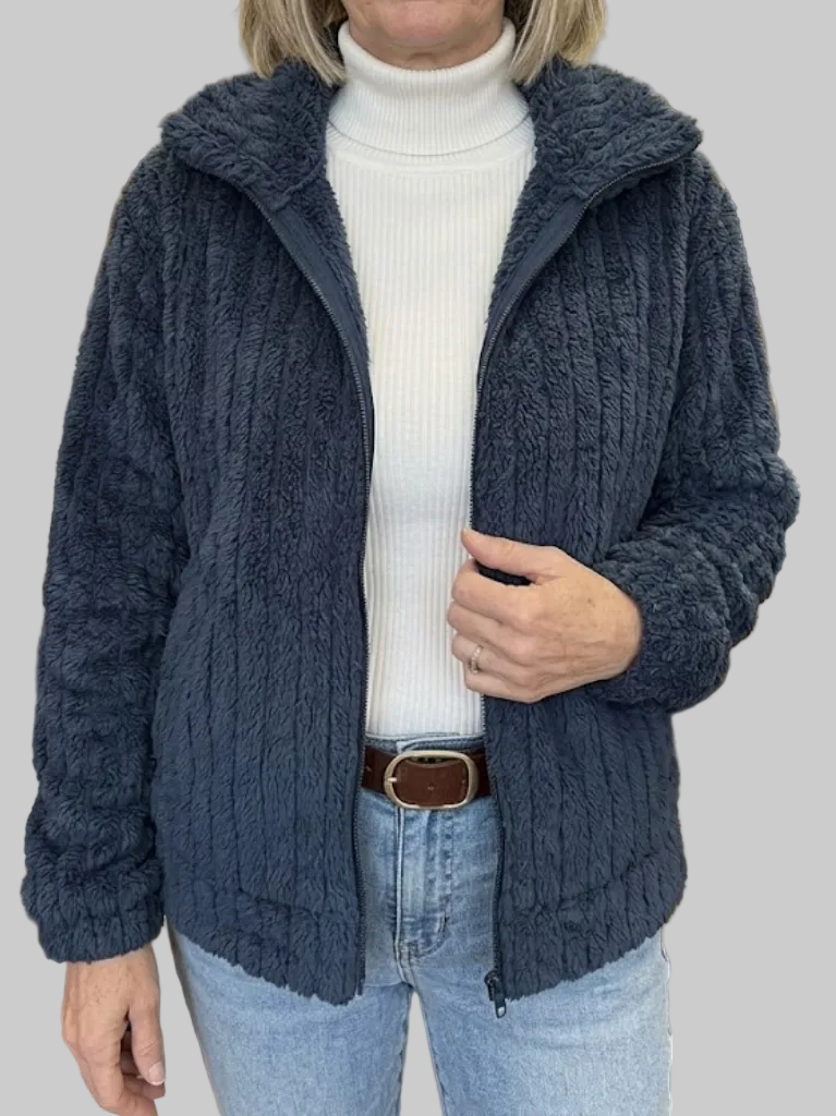 A woman wearing an open dark navy blue ribbed fleece jacket with a fold-over collar, styled over a white ribbed turtleneck top and blue jeans.