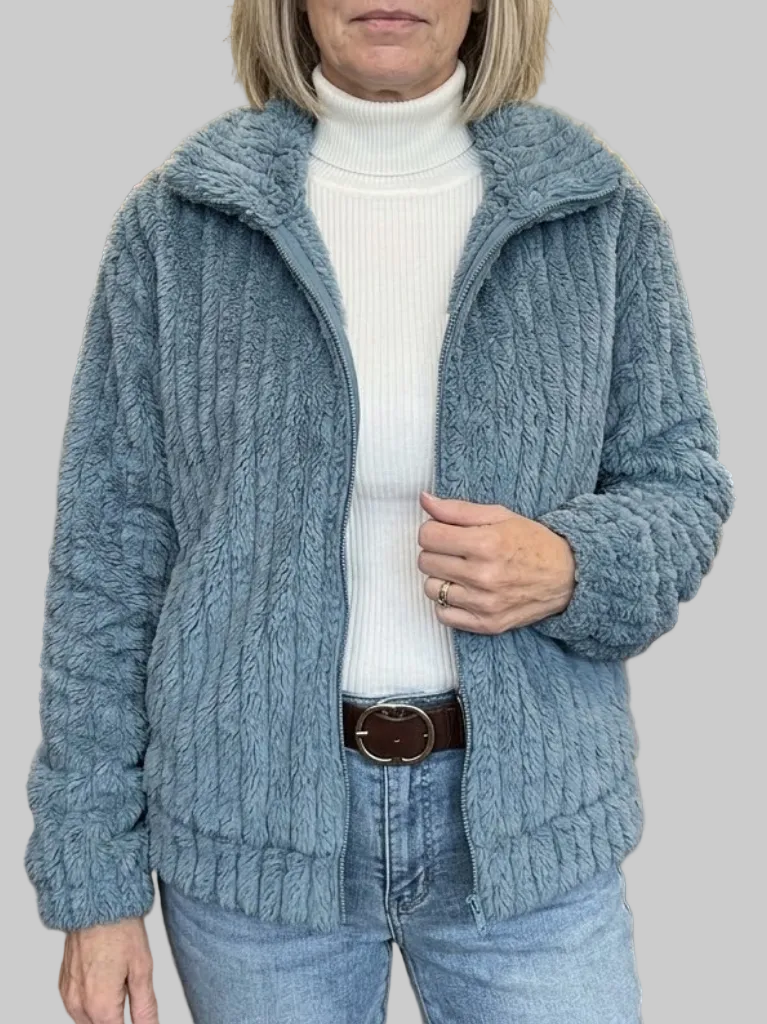 A woman wearing an open slate-blue ribbed fleece zip-up jacket with a spread fold-over collar, layered over a white turtleneck and blue jeans.