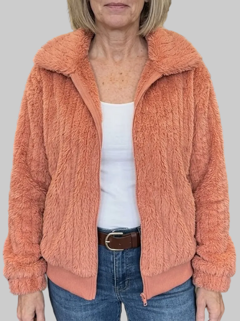 A woman wearing an open bright coral-orange ribbed fleece jacket with a fold-over collar, layered over a white top and blue jeans.