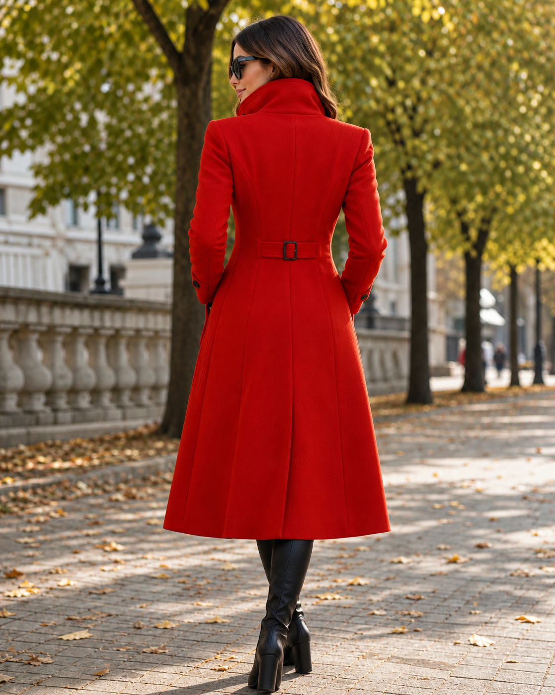 Rear full-length shot of a woman wearing a tailored red longline wool coat showing the flared A-line hem and back waist belt tab detail on an outdoor path.