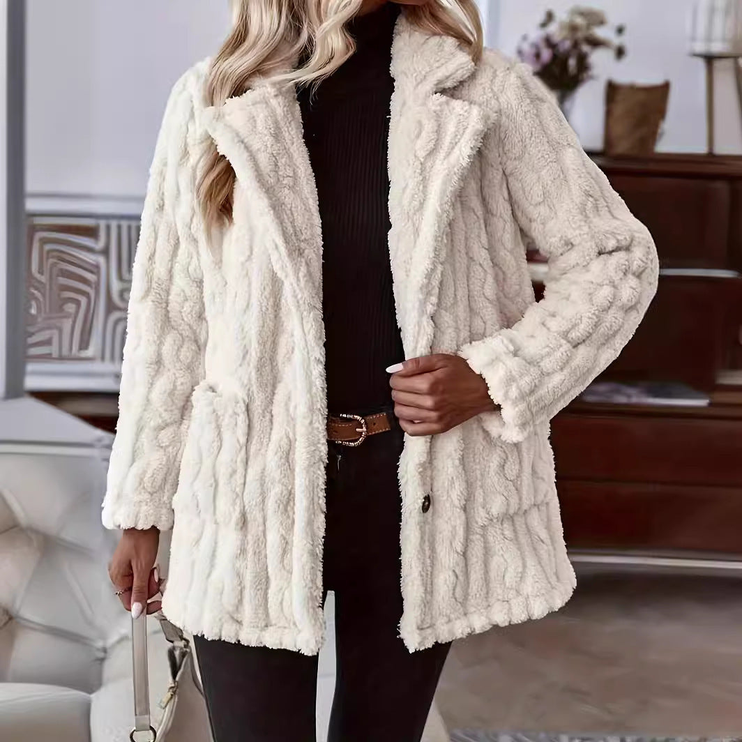 A woman with blonde wavy hair wearing an off-white cream teddy fleece coat with an embossed cable-knit pattern, wide notch lapels, front patch pockets, and button closure, styled open over a black turtleneck top and dark jeans with a brown belt.