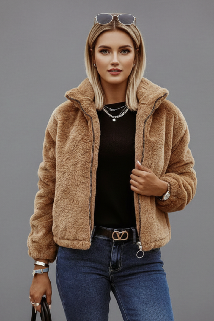 A blonde model wearing a tan teddy fleece jacket with a stand collar, styled open over a black top, dark blue jeans, and sunglasses.