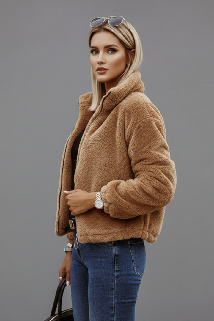 Side profile of a model wearing a tan teddy fleece zip-up jacket with a stand collar paired with blue jeans.
