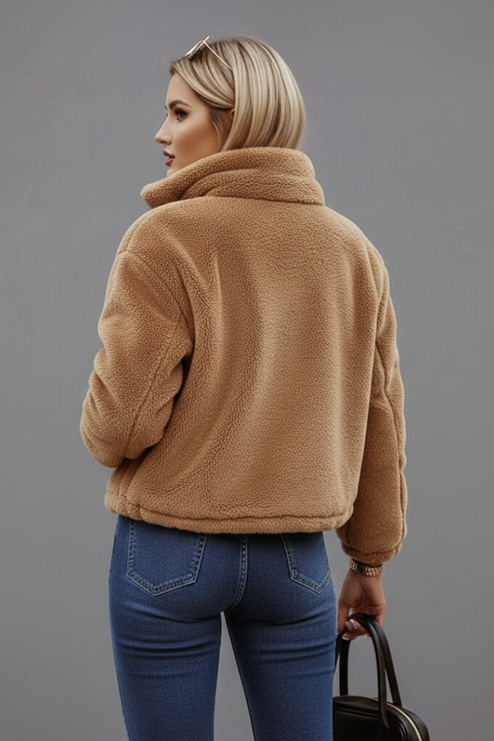 Back view of a model wearing a warm brown teddy fleece jacket and blue jeans, holding a black handbag.