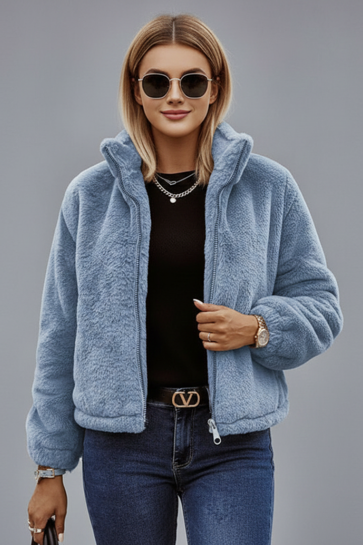 A model wearing a dusty light blue teddy fleece zip-front jacket styled with round sunglasses, a black top, and jeans.