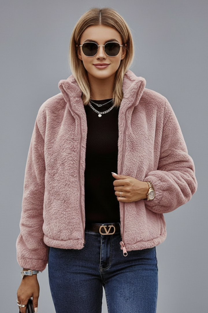 A model wearing a dusty rose pink teddy fleece jacket with a stand collar, styled over a black shirt and dark blue jeans.