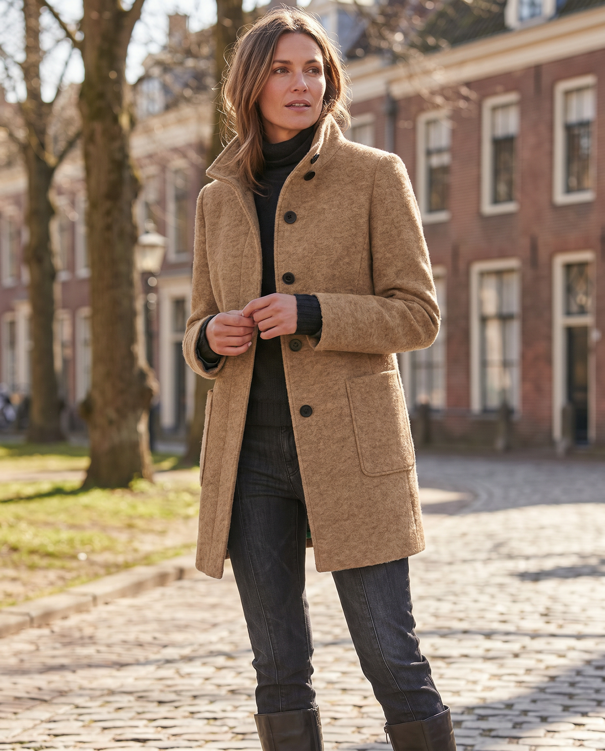 Full-length outdoor view of a model walking down a cobblestone street wearing the camel wool stand-collar coat.