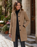 Full-length street style portrait of a model wearing a camel brown women's wool coat featuring a straight fit silhouette, high stand collar, black button front, and large patch pockets, layered over a black turtleneck and black pants.