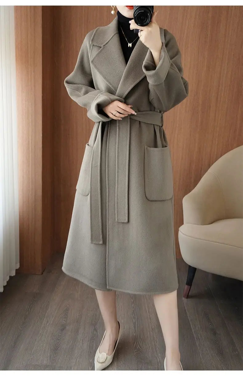 Full-length mirror selfie of a woman wearing a light greyish-sage longline wool-blend coat tied at the waist with a fabric belt and paired with beige flats[cite: 10].