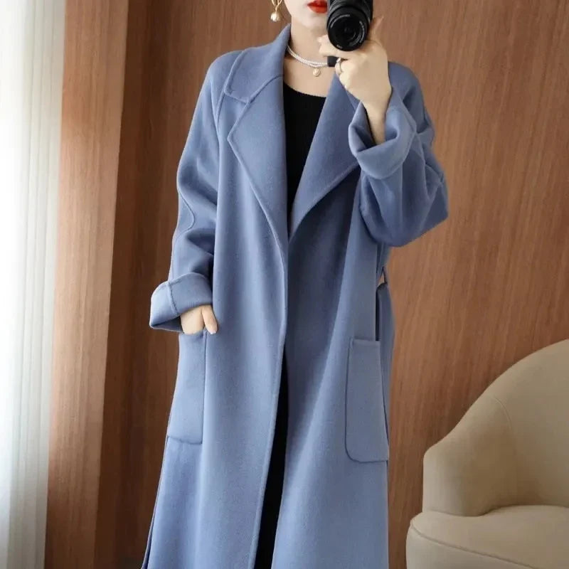 Full-length mirror selfie of a woman wearing a dusty blue longline wool-blend overcoat featuring wide lapels and front patch pockets.
