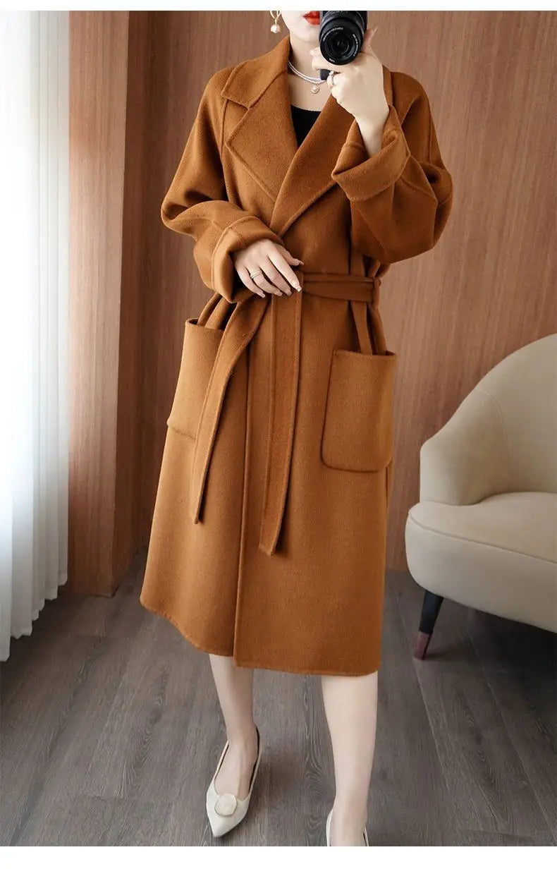 Full-length mirror selfie of a woman wearing a caramel brown longline wool-blend coat styled with a tied belt at the waist and large front patch pockets
