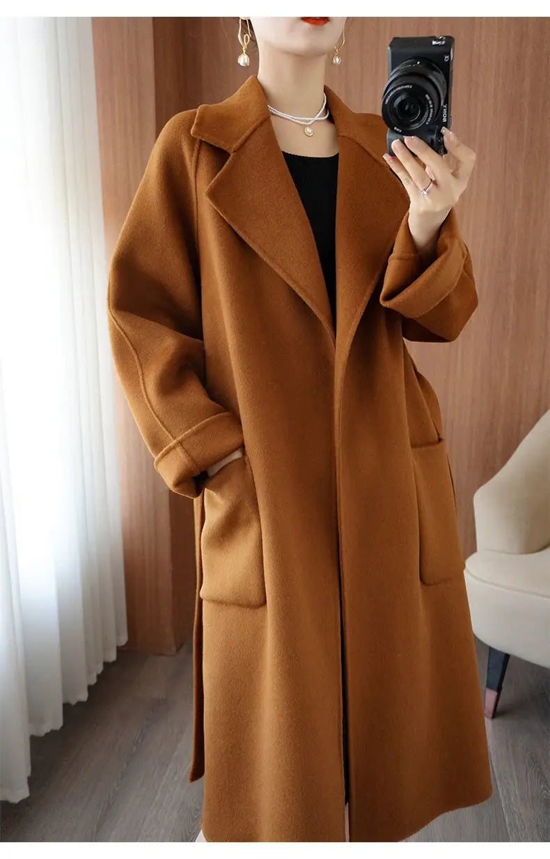 Three-quarter angle mirror selfie of a woman wearing an unbelted caramel brown wool-blend longline overcoat with wide sleeves.