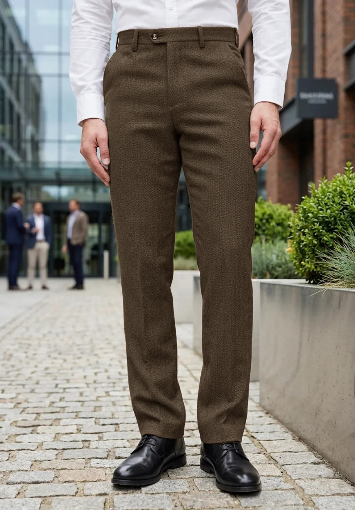 Full-length front view of a man wearing brown wool herringbone straight-leg dress pants, a white dress shirt, and black shoes standing on a paved outdoor walkway near modern city buildings.