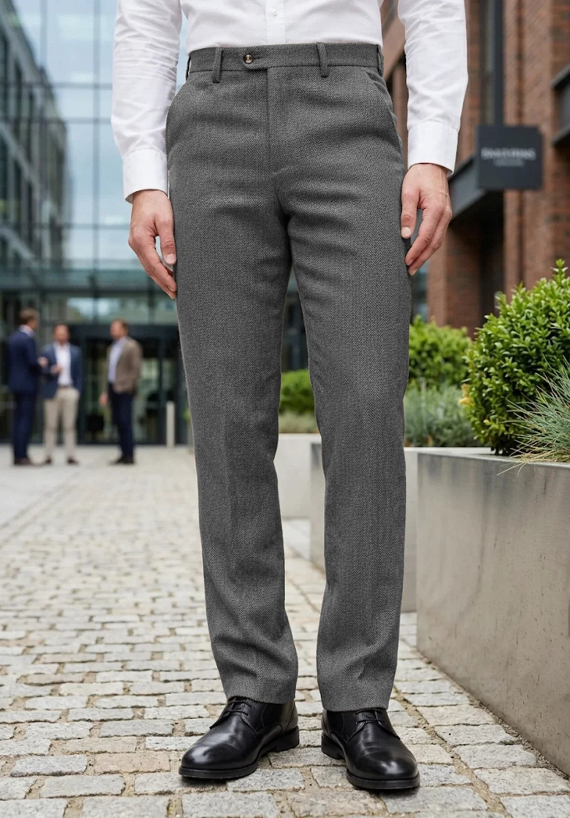 Full-length front view of a man wearing gray wool herringbone dress pants, a white dress shirt, and black shoes standing on an outdoor city sidewalk.