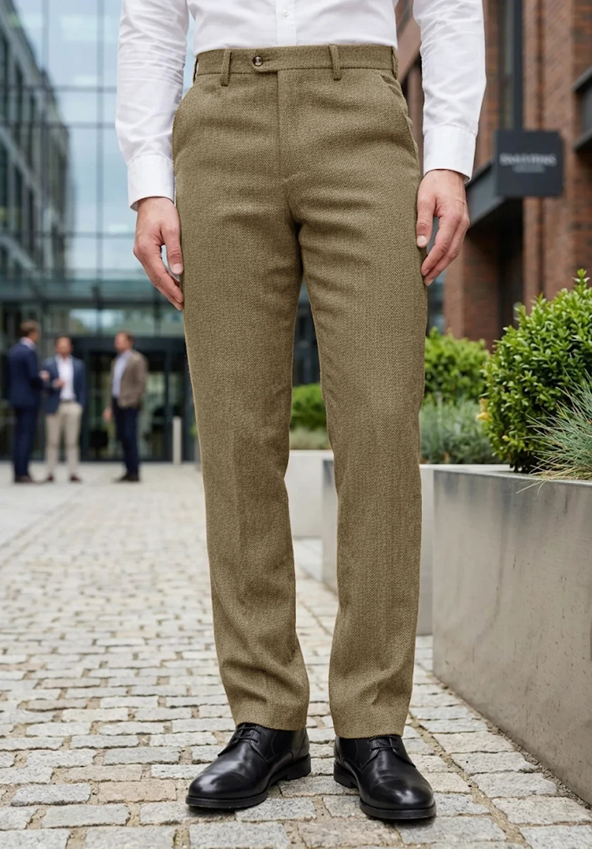 Full-length front view of a man wearing olive green wool herringbone dress pants, a white dress shirt, and black dress shoes standing outdoors in an urban setting.