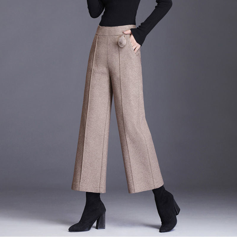 Front view of a model wearing high-rise beige wool wide-leg trousers with front center seams, side pockets, and a plush pom-pom accent, paired with black pointed boots.