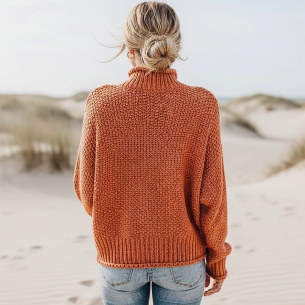 Rust orange chunky knit turtleneck sweater with an oversized fit and ribbed trim, worn outdoors at a sandy beach.