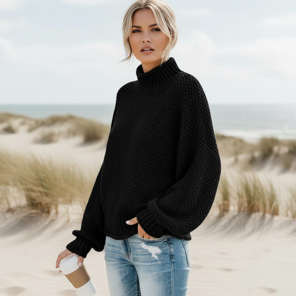 Black chunky knit turtleneck sweater with balloon sleeves and an oversized fit, worn with denim on a sandy beach.