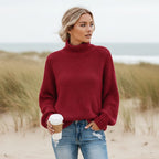Burgundy chunky knit turtleneck sweater with balloon sleeves and an oversized fit, worn with distressed denim on a beach.