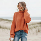 Rust orange chunky knit turtleneck sweater with balloon sleeves and an oversized fit, worn with denim on a beach.