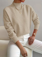 Beige ribbed turtleneck sweater with dropped shoulders and fitted cuffs, styled with white trousers.