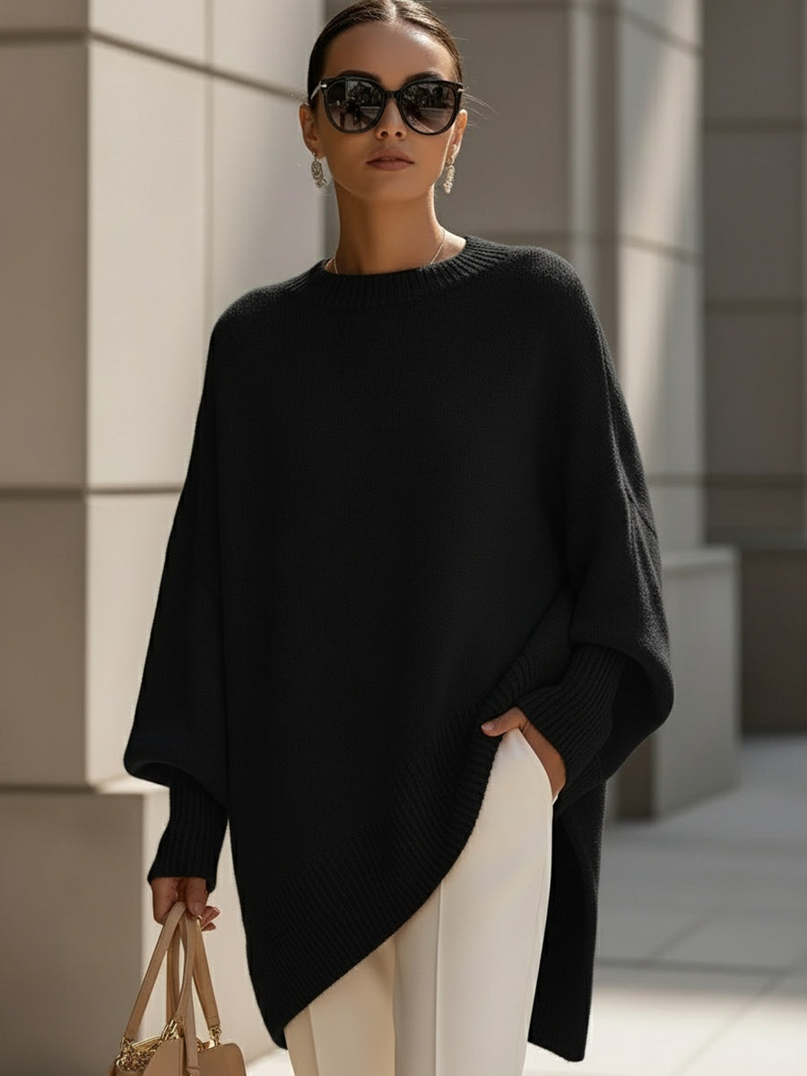 Black oversized ribbed knit sweater with a loose poncho silhouette, long sleeves, and structured cuffs worn with trousers.