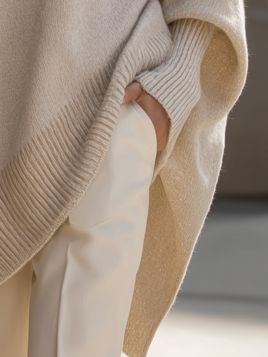 Beige ribbed knit oversized poncho sweater with structured cuffs worn over cream trousers.