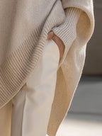 Beige ribbed knit oversized poncho sweater with structured cuffs worn over cream trousers.