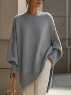 Grey oversized ribbed knit sweater with a relaxed poncho silhouette and structured cuffs worn with cream trousers.