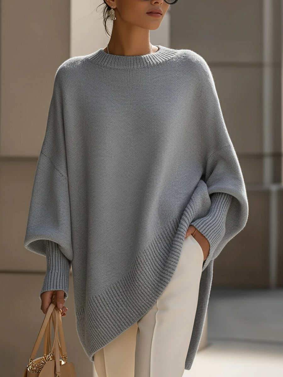 Grey oversized ribbed knit sweater with a relaxed poncho silhouette and structured cuffs worn with cream trousers.